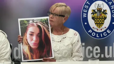 A woman who has short blonde hair and is wearing a white top is holding up a large poster of a head-and-shoulder image of a woman, Reanne Coulson, who has long brown hair. In the background is a logo of the West Midlands Police. 