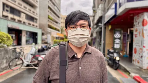 BBC/Tessa Wong Man with salt and pepper hair wearing wire glasses and face mask posing for camera. He was wearing a brown polo shirt and you could see the belt on his bag on his shoulders. In the background, you can see the streets of Taipei, including parked bicycles and motorcycles.