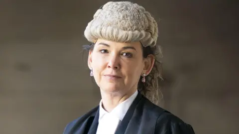 Dorothy Bain, who has long dark hair tied back under a court wig, looks straight at the camera. 