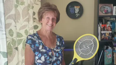 BBC Lady with blue top and electric fly swatter in a house setting 