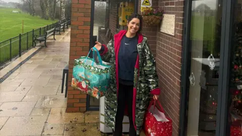 A woman dress in black with a long camoflage coat is standing outside of a building holding two large Christmas bags