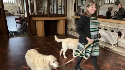 Woman wearing a green, black and grey striped dress with a black cardigan, black tights and shoes and leading two dogs through a church