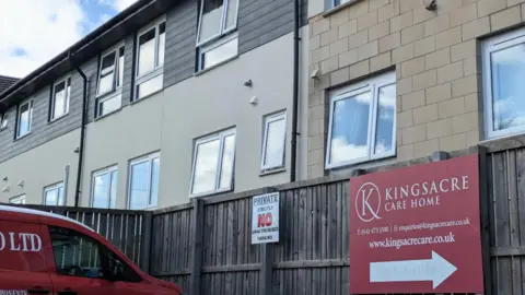 Exterior view of a multi-storey care home building behind a wooden fence, with a red sign reading “Kingsacre Care Home” and a red van parked in the foreground. Several windows are visible across the cream and grey-clad façade, along with a private parking notice on the fence.