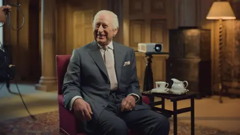 Tim Cragg/Passion Planet/PA Wire King Charles laughing while sitting on a chair in front of a projector in a wood-panelled room. 