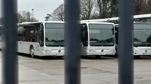 A row of silver buses parked at the bus depot in Douglas. the bars of a black metal fence are in the foreground.
