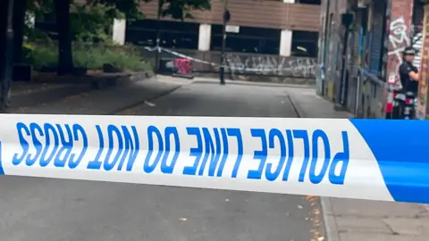 A blue and white stretch of police tape with the words 'police line do not cross' printed in blue letters. In the background is a short stretch of road flanked by commercial premises with a female police officer in a black uniform stood to the right of the frame. 