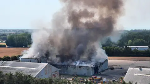 A metal warehouse with a plume of smoke coming from the roof. Fire engines can be seen parked next to the building.