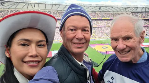 Andrew Wills Three people in the foreground pose for a photograph in a large sports stadium. One person wears a red-and-white hat, another wears a striped knitted hat and padded gilet, and a third wears a Bristol Bears rugby shirt. A rugby pitch is visible behind them, with large flags laid out on the grass and stadium seating filled with spectators.