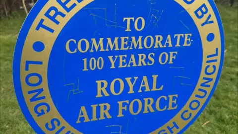 A blue circular sign with gold writing on. Its a plaque commemorating 100 years of the Royal Air Force. Nazi Swastikas have been engraved onto it. 