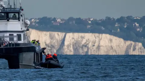 BBC A small boat containing migrants in the English Channel off the coast of Dover receiving support from a larger Border Force boat. The white cliffs are in the background