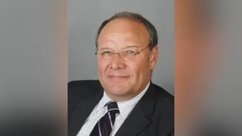 Stuart Munro's official councillor photo, with him dressed in a suit looking at the camera. He has short, grey hair and is wearing glasses.