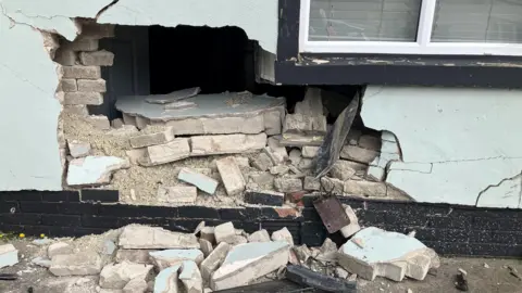 To the left hand side of the a broad living room window, on the external wall of a house, there is a hole in the wall through which you can see the inside of the house. Broken bricks have spilled onto the driveway.