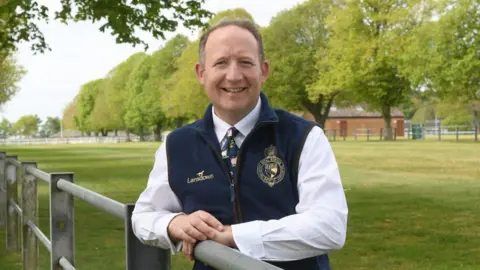 Mark Nicholas leans on a metal gate. Around him are lots of trees and green grass. Mark is wearing a white shirt, blue tie and blue gilet. He has short grey/brown hair.