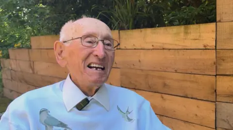 George King/BBC An elderly man wearing a blue jumper with birds on it over a white shirt. He is wearing glasses and sitting next to a wooden wall. He is smiling.
