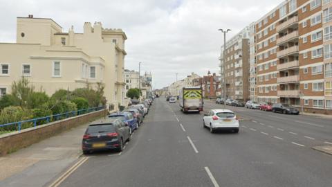 A street-view image of a two-lane carriageway with houses on the left side and blocks of flats on the right