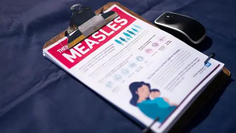 A fact sheet for measles sits on a table with a dark blue tablecloth, in a clipboard