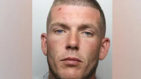 A custody photo shows a man with a shaved head and blue eyes and a cut on his forehead. He is wearing a grey t-shirt.