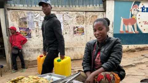 Sami Awami/BBC A woman in a black jacket sells food at a street stall. Behind her walks a man wearing a baseball cap and carrying yellow cans.