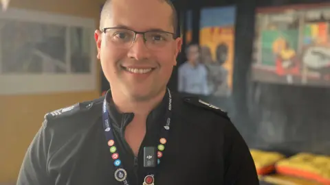 Jon Wright/BBC Jason Thomas, in police uniform, looks at the camera. There is artwork behind him.