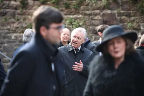 Athena A man in a black jacket and tie and white hair is standing speaking