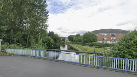The view from on top of a bridge. The view shows a blue and white metal fence running along the road. Below the bridge is a narrow canal with trees either side of it. A grass bank with railings running along the canal can be seen on the right-hand side. A series of-three storey flats are also positioned on the right. 