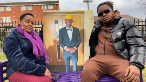 Irene and Elliot sitting together on a purple bench dedicated to Eddie at the park. Irene is wearing a long burgundy skirt, a soft blue jacket and a purple scarf. Elliot is wearing sunglasses and a full brown tracksuit under a padded black shiny raincoat. In between them is a big picture of Eddie wearing a suit, green bow tie and a yellow cap, holding a glass of champagne and smiling at the camera.