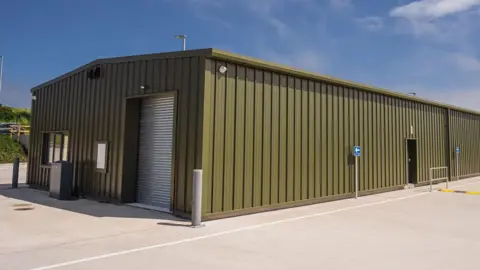 ECAS A large khaki metal shed with corrugated door at the front within a large concrete compound. 