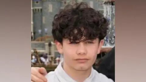 A young boy wearing a white polo shirt. He has curly brown hair. Someone has their hand on his shoulder. Behind, out of focus in the background, is a medieval castle-looking building.