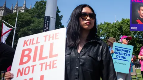 A woman in sunglasses holds a sign that says "Kill the bill" in capitals. She is surrounded by other protesters, one of whom is holding a sign that says "support assisted dying".