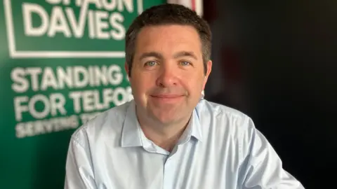 A dark-haired man wearing a pale blue shirt sitting in front of a blurred green board and black wall