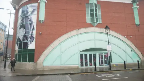 Exterior shot of the former Debenhams store in St Marks. It has a curved green-coloured panel over the glass doors, A person is walking on the path nearby.