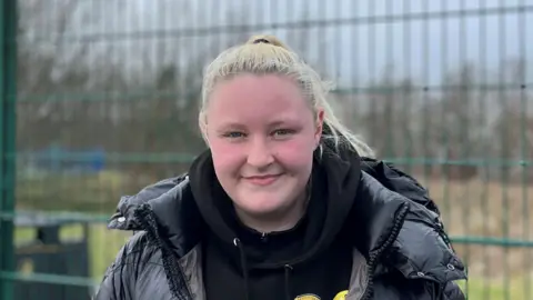 Teenager Jodie has long, blonde hair pulled back into a high ponytail. She is standing outside, next to a metal fence, and is wearing a black hoodie and a black padded jacket. She is smiling at the camera.
