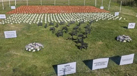 Northamptonshire Police A row of black silhouettes, flowers, white doves and flags, all in rows on a piece of grass. There are signs in front of the display.