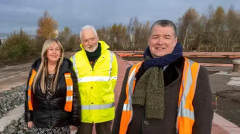 National Lottery Heritage Fund Deirdre Gaughan, Andy Milne and Councillor Ricky Bell stand smiling at a building site. There are trees behind them but they stand in rubble. You can see the transport museum in the background. 