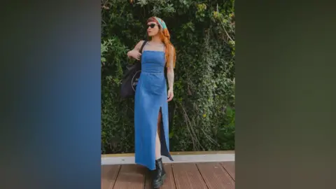 Sophie Saint Sophie is posing in a long denim strapless dress with black Doc Martens, black sunglasses, a russet and turquoise-coloured headscarf and large black bag. She has auburn hair. She is standing on decking with greenery behind.