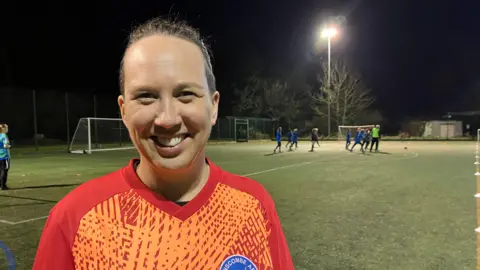 BBC A women is smiling at the camera. She is standing on a football pitch with players in the background. The pitch is lit with floodlights. She has brown hair tied back and is wearing an orange patterned football top. 