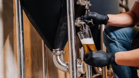 A person holding a glass and pouring a beer from a large vessel.