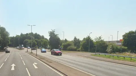 The A602, showing a dual-carriageway with cars on the road and a supermarket to the right. There is a junction with road markings on it. Trees are by all the entrances. 