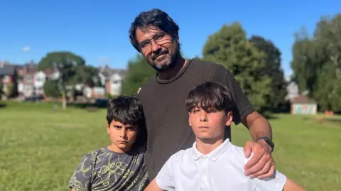 Hugo in a dark olive T-shirt with his arms around his sons Luca, in a grey and yellow patterned T-shirt, and Guilherme in a white polo shirt, in a park in Exeter.