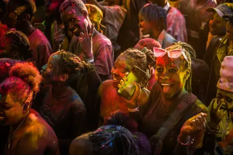 Gerald Anderson / Getty Images Crowds of smiling people are covered in multicoloured powder.