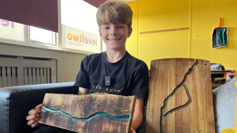 Jon Wright/BBC Oscar, 10, sits holding a wooden board with a blue resin filled groove across it.