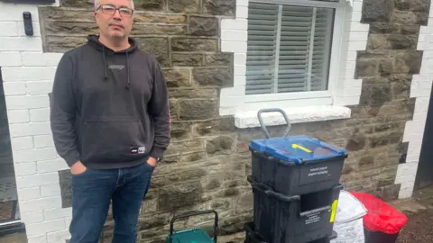 BBC Mark Underdown who is wearing a black hoodie and blue jeans with glasses and his hands in his pockets, stood outside a house with a stack of bins beside him