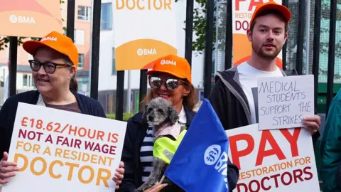 PA Media NHS resident doctors protest outside Royal Victoria Infirmary in Newcastle. They are holding plaquards and wearing orange caps that say 'BMA' 