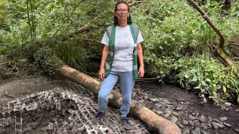 Simon Thake/BBC A woman in a green jacket and blue trousers stands next to a stream in woodlands. 
