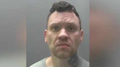 Cambridgeshire Police Paul King's police custody image. He looks at the camera with his mouth corners slightly pointing down. He wears a grey top and has a neck tattoo.