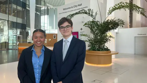 An image of a teenage girl and boy smartly dressed in the City Hall. A large shrubbery sites on their right.