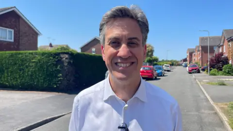 Ed Miliband wearing a white shirt smiles at the camera while standing on a residential street on a sunny day.