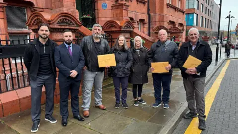 Astley Warehouses Action Group A group of people stand on a street holding letters. 