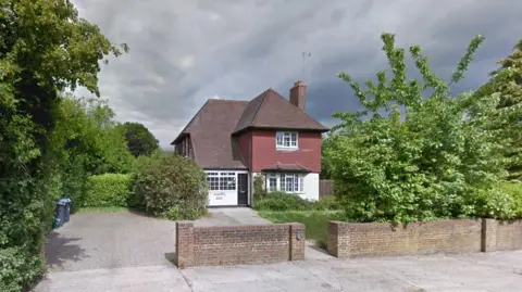 The outside of a detached house which has a white rendered lower half and red tiled about. There is a driveway and a front lawn with shrubs growing around the edge as well as trees with green leaves behind a low brick wall