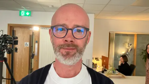 A bald man with a grey beard, wearing glasses, a white t-shirt and navy cardigan.